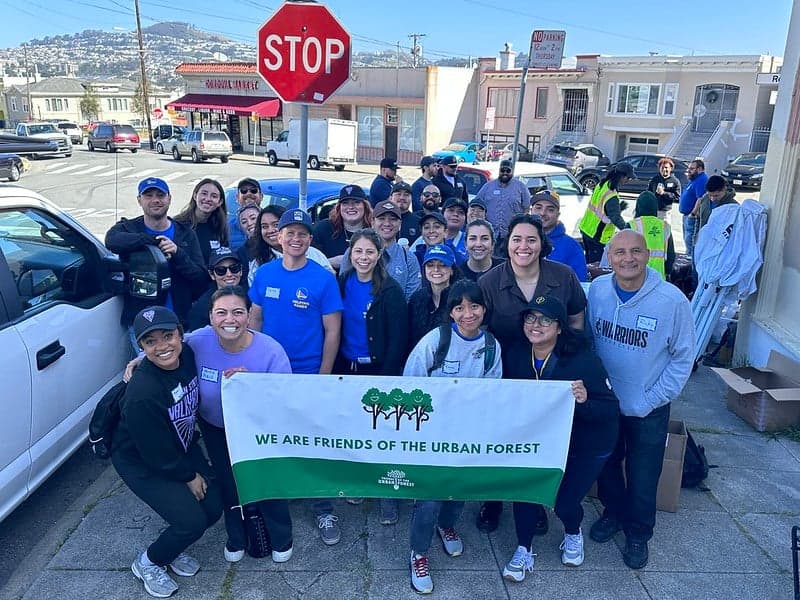 volunteers huddled on planting day, holding our sign featuring the Friends of the Urban Forest logo and happy trees.